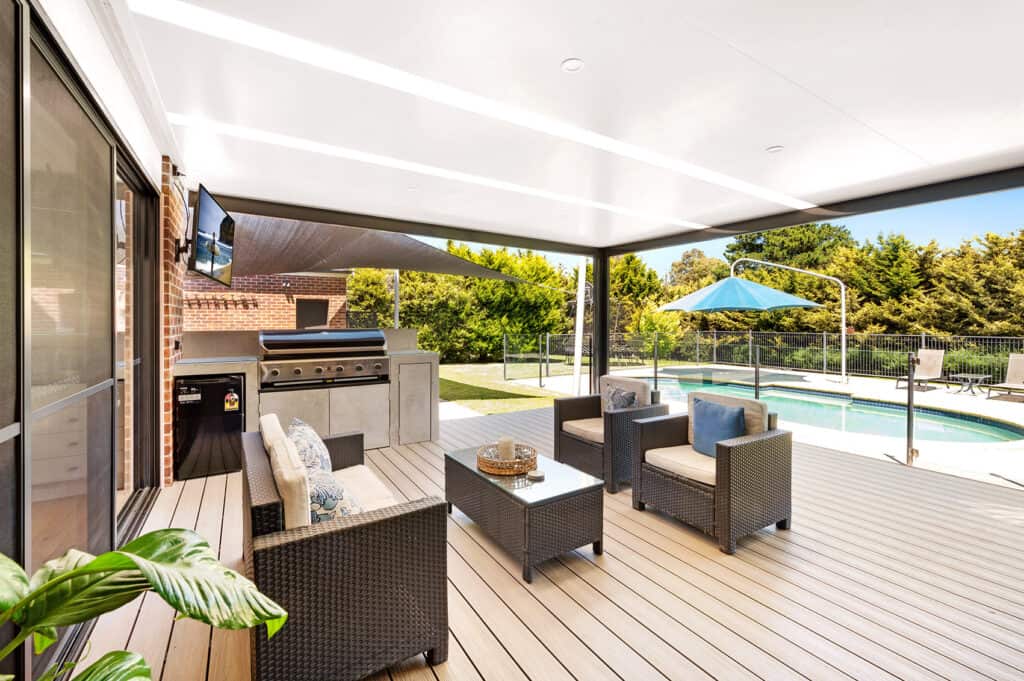 A flat roof patio roof covering a seating and BBQ outdoor living space with a pool in the background.